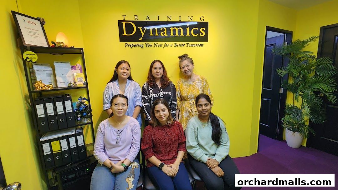 A group of six women posing in front of the Training Dynamics logo and shelves.