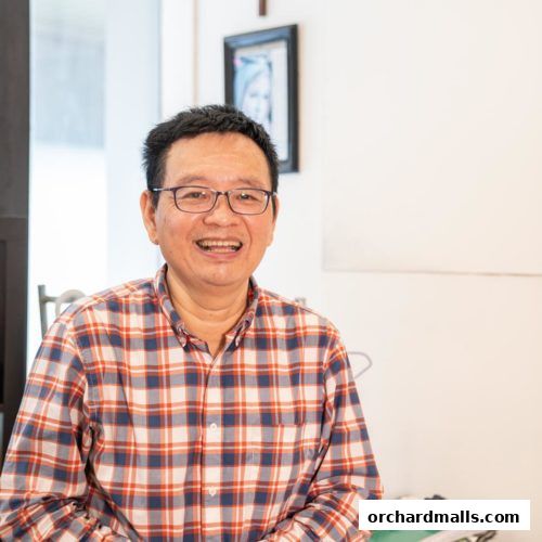 A smiling man in a plaid shirt wearing glasses, with a framed picture behind him.