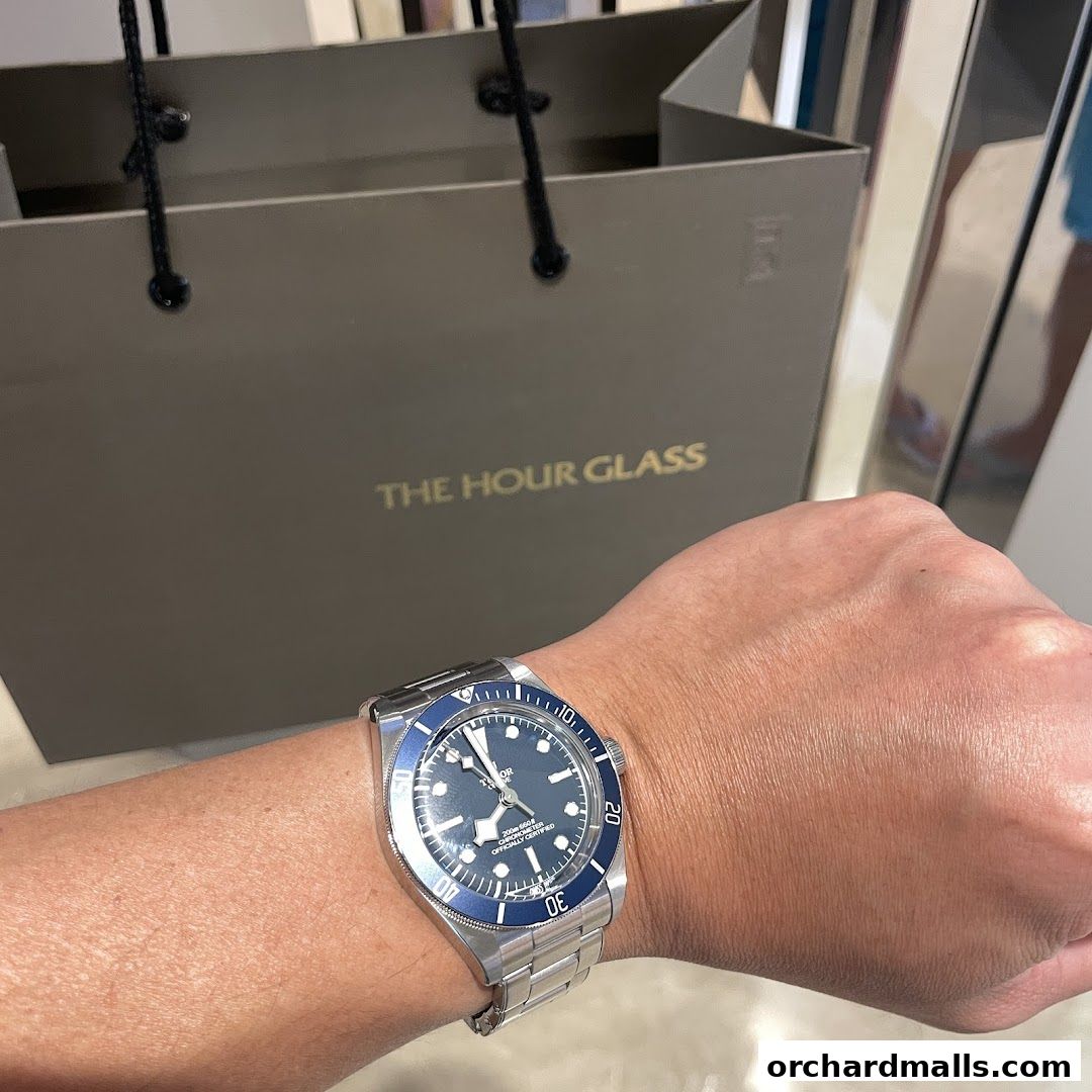 A person wearing a Tudor watch with a shopping bag from The Hour Glass in the background.