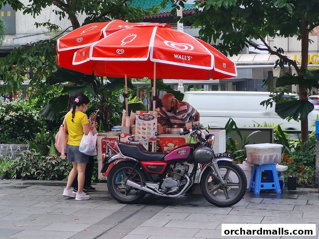 An ice cream cart with a red umbrella and a motorcycle parked nearby.
