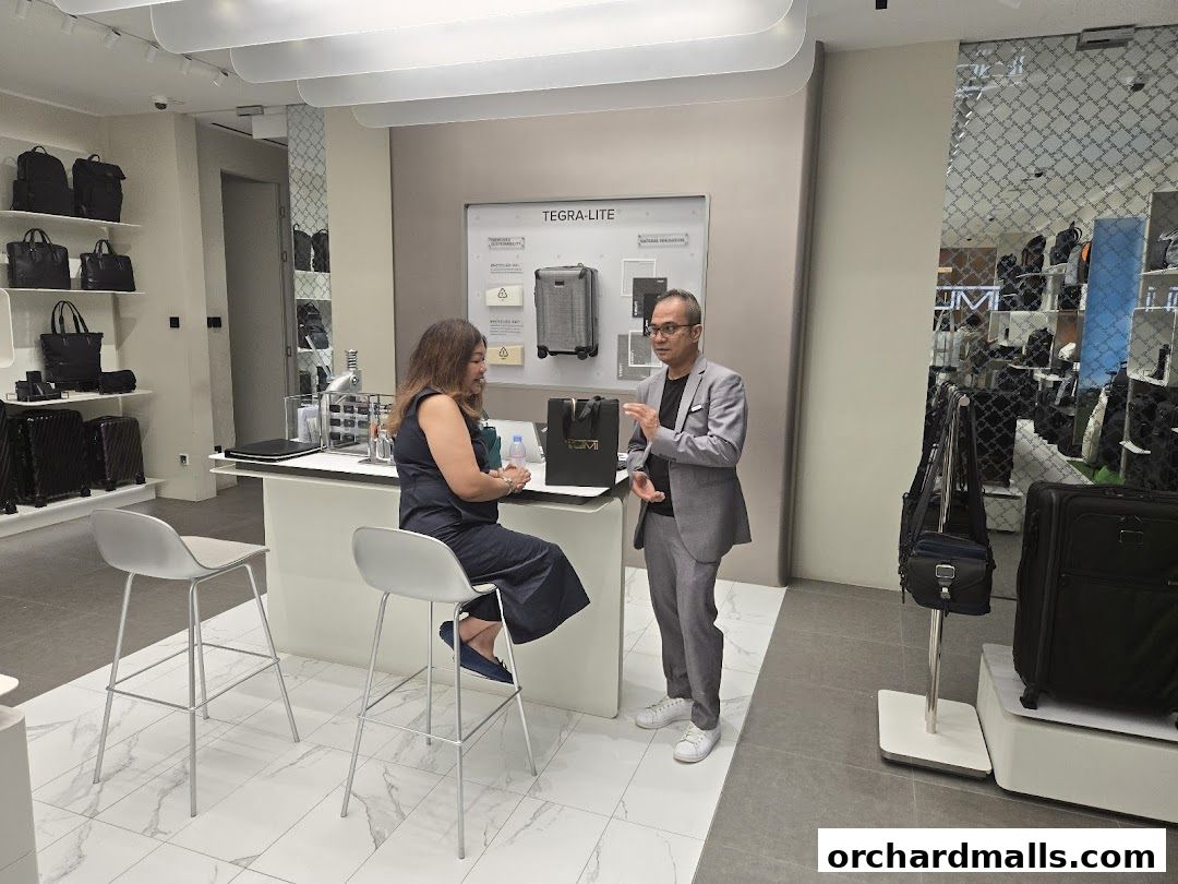 A man in a suit explains TUMI products to a seated woman in a store.