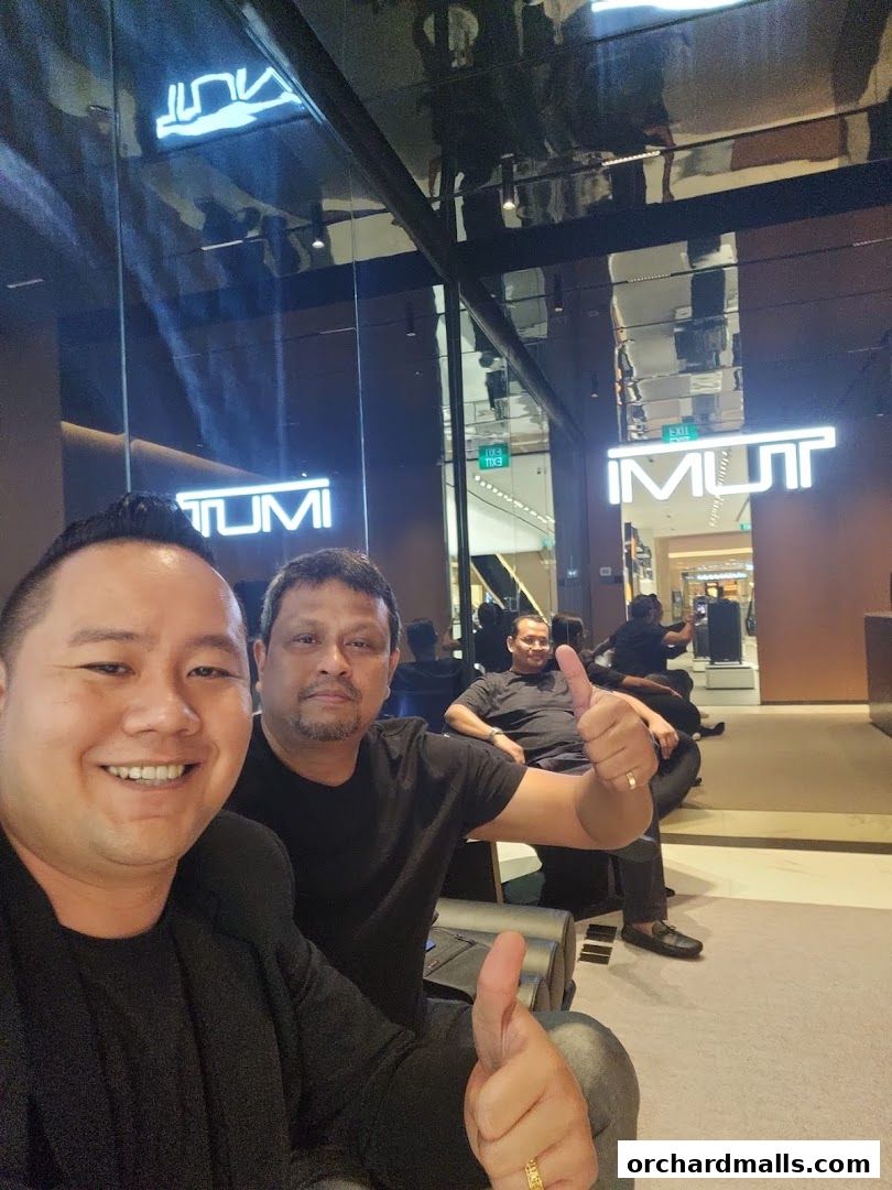 Two men give a thumbs up in front of a TUMI store with illuminated signage.