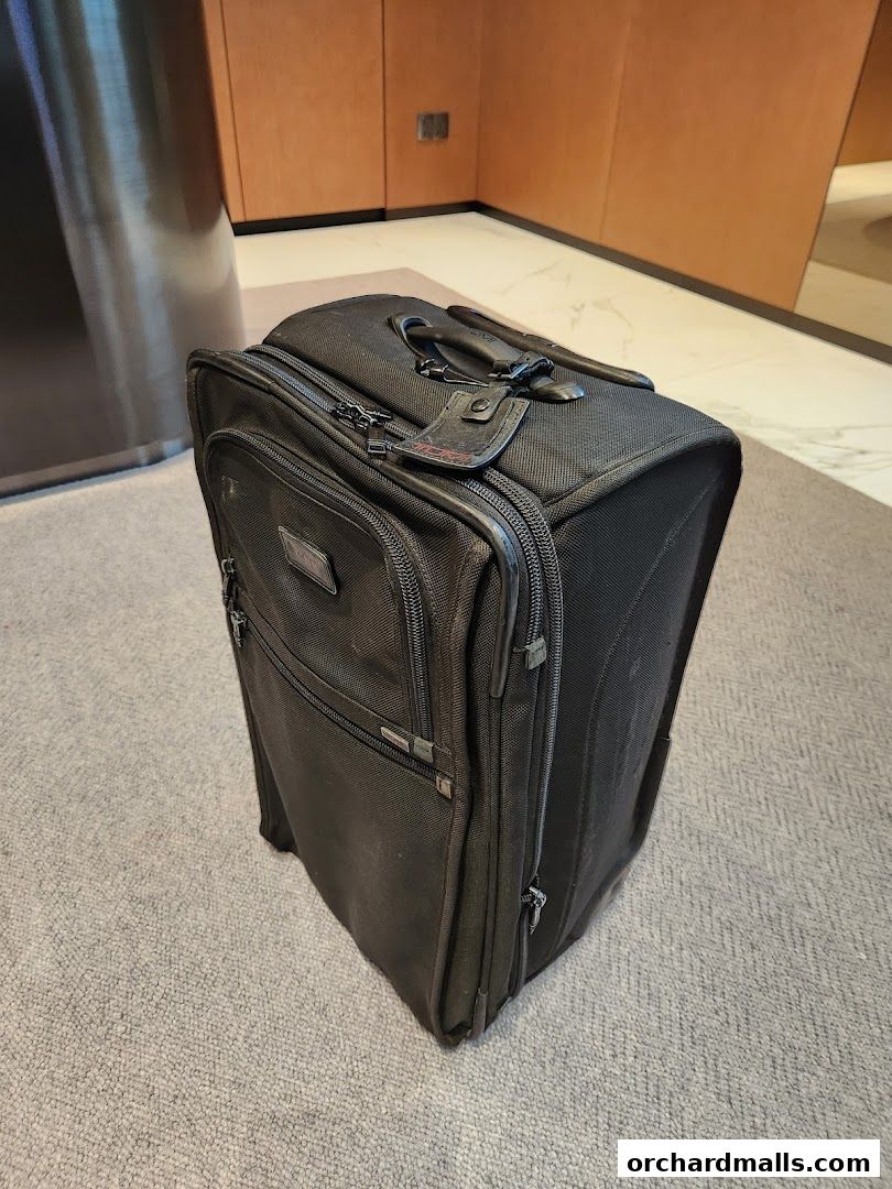A black TUMI suitcase with a telescopic handle and luggage tag sits on a carpeted floor.