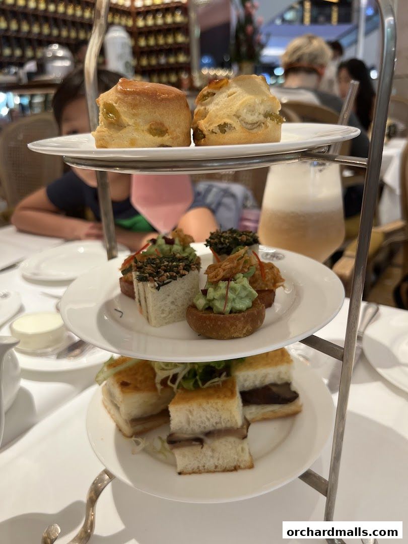 A tiered stand displays a selection of delicate pastries and sandwiches, with people dining in the background.