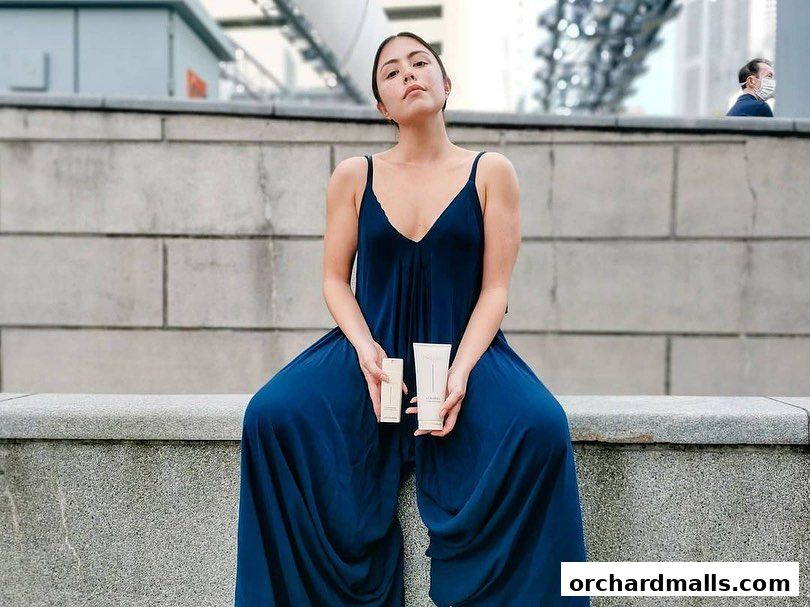 A woman in a blue jumpsuit holds two skincare products while sitting outdoors.
