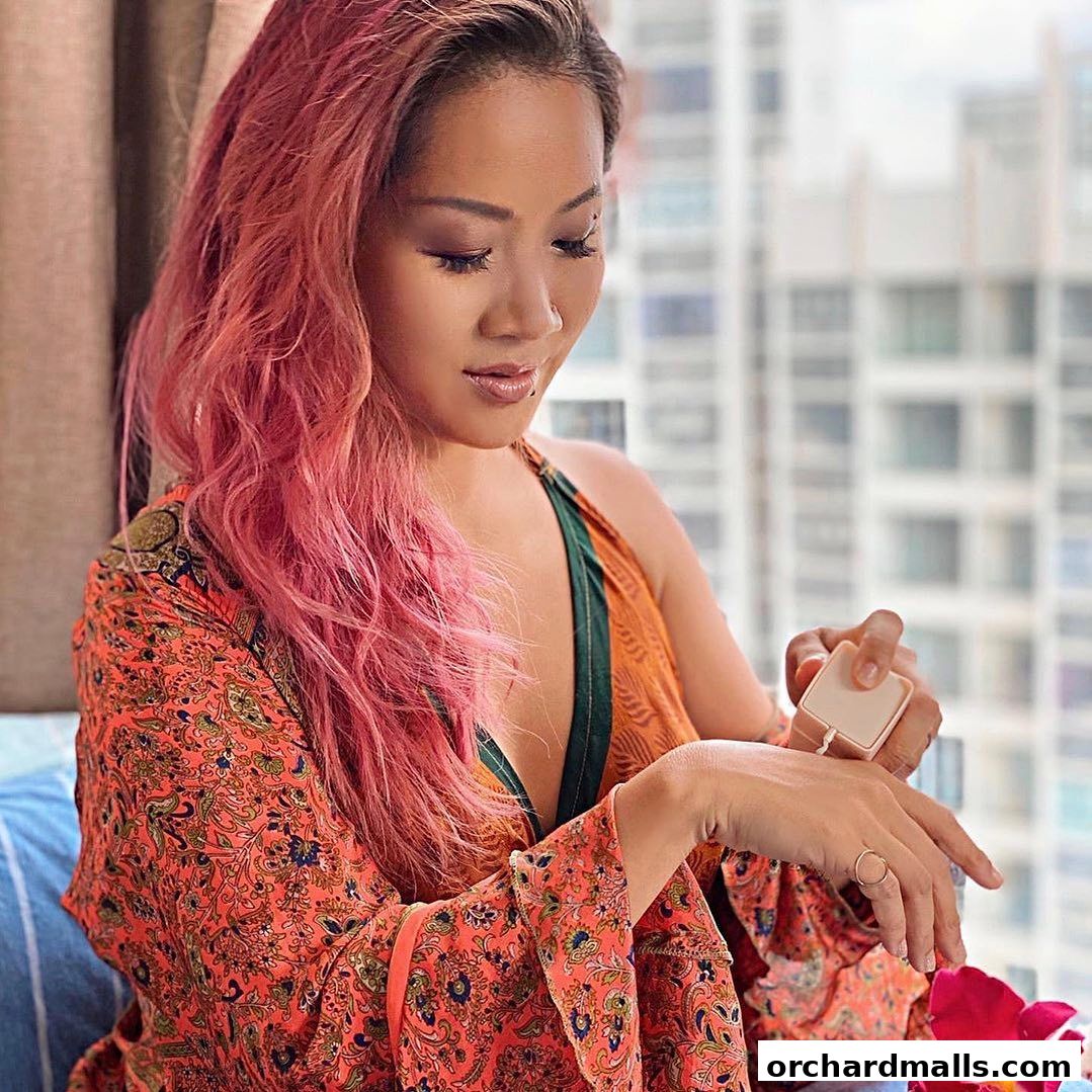 A woman with pink hair applies a product to her hand, with a city skyline in the background.