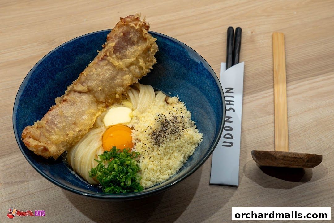 A bowl of udon noodles with tempura, egg yolk, and chives, served with chopsticks.