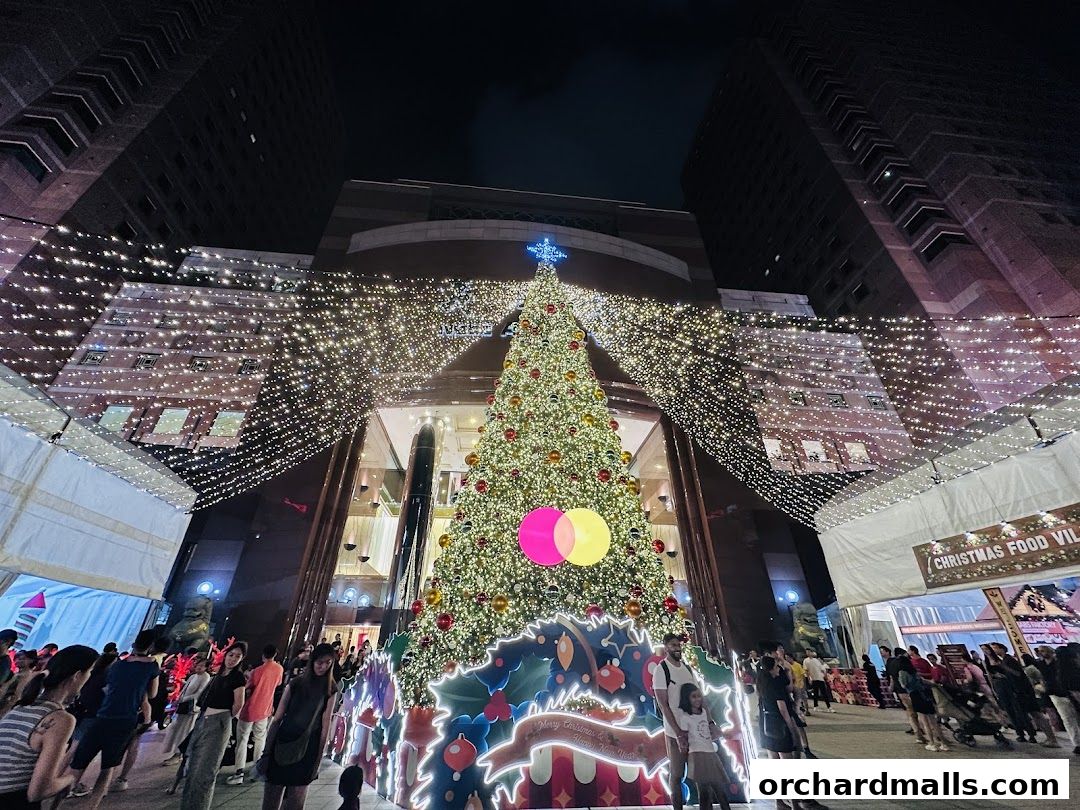 A festive Christmas tree and lights adorn a shopping mall entrance with people milling about.