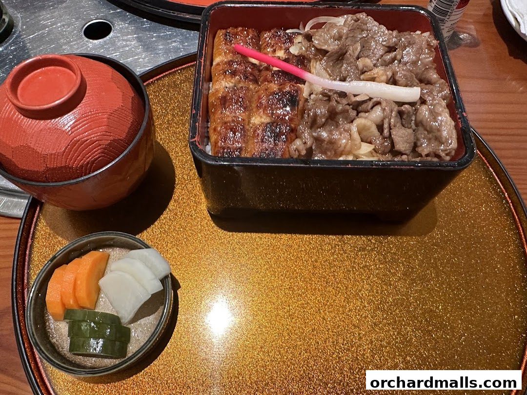 A bento box with grilled eel and beef over rice, served with pickles and a soup bowl.