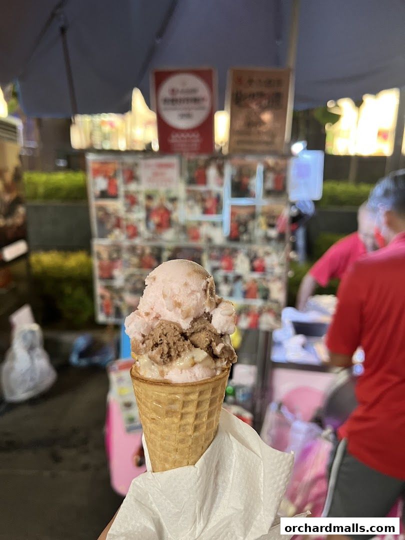 A hand holds a waffle cone filled with three scoops of ice cream, with a shop in the background.