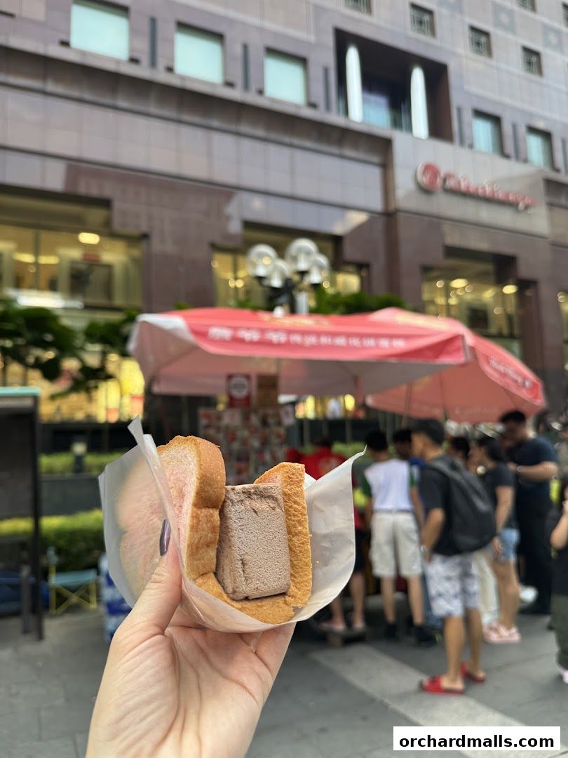 A hand holds a traditional wafer ice cream sandwich in front of a bustling street stall.