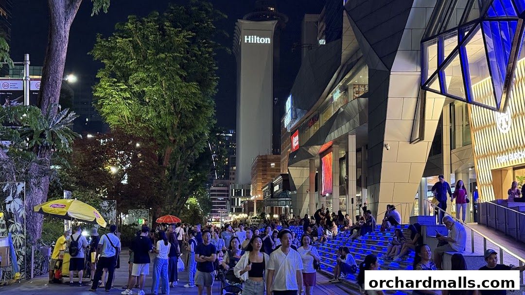 A lively street scene at night with many people walking and sitting, illuminated by building lights and a prominent Hilton tower.