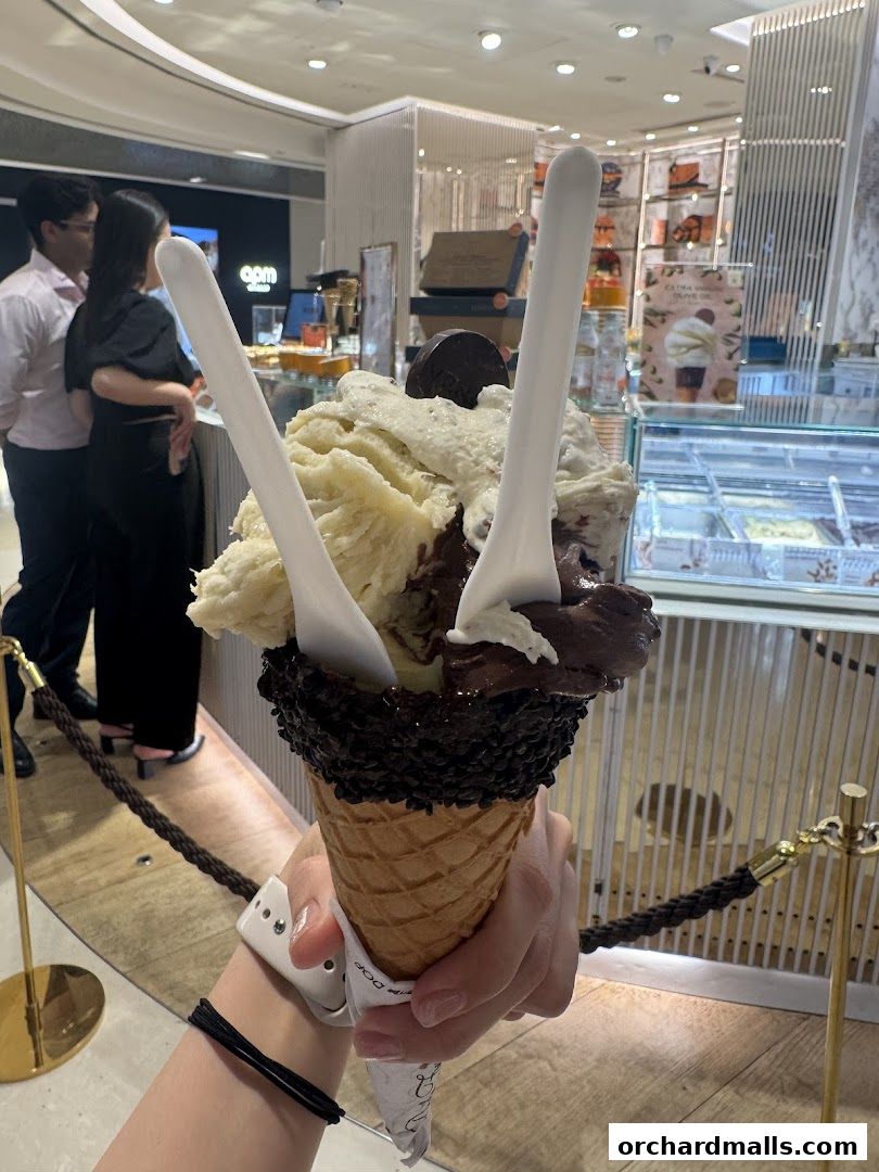 A hand holds a large gelato cone with two flavors and chocolate sprinkles in front of a store.