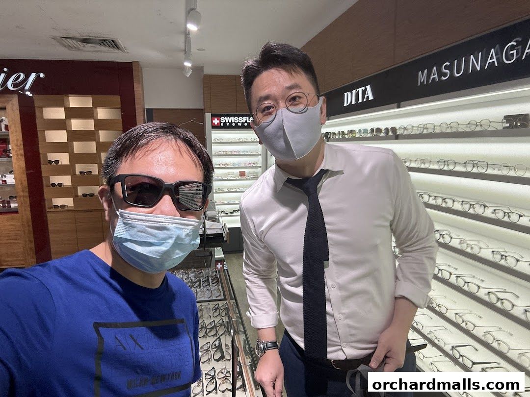 Two men in masks stand in an optical shop, surrounded by displays of eyeglasses.