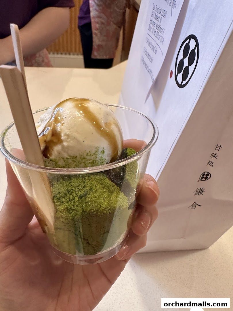 A hand holds a clear cup filled with matcha mochi, ice cream, and syrup.