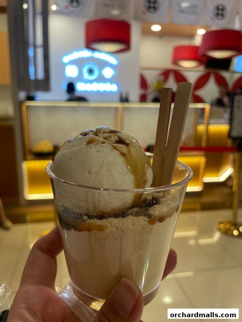 A hand holds a clear cup filled with ice cream, sauce, and powder.