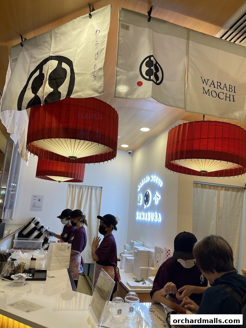 Staff serving customers at a Warabi Mochi Kamakura shop with decorative banners and lamps.