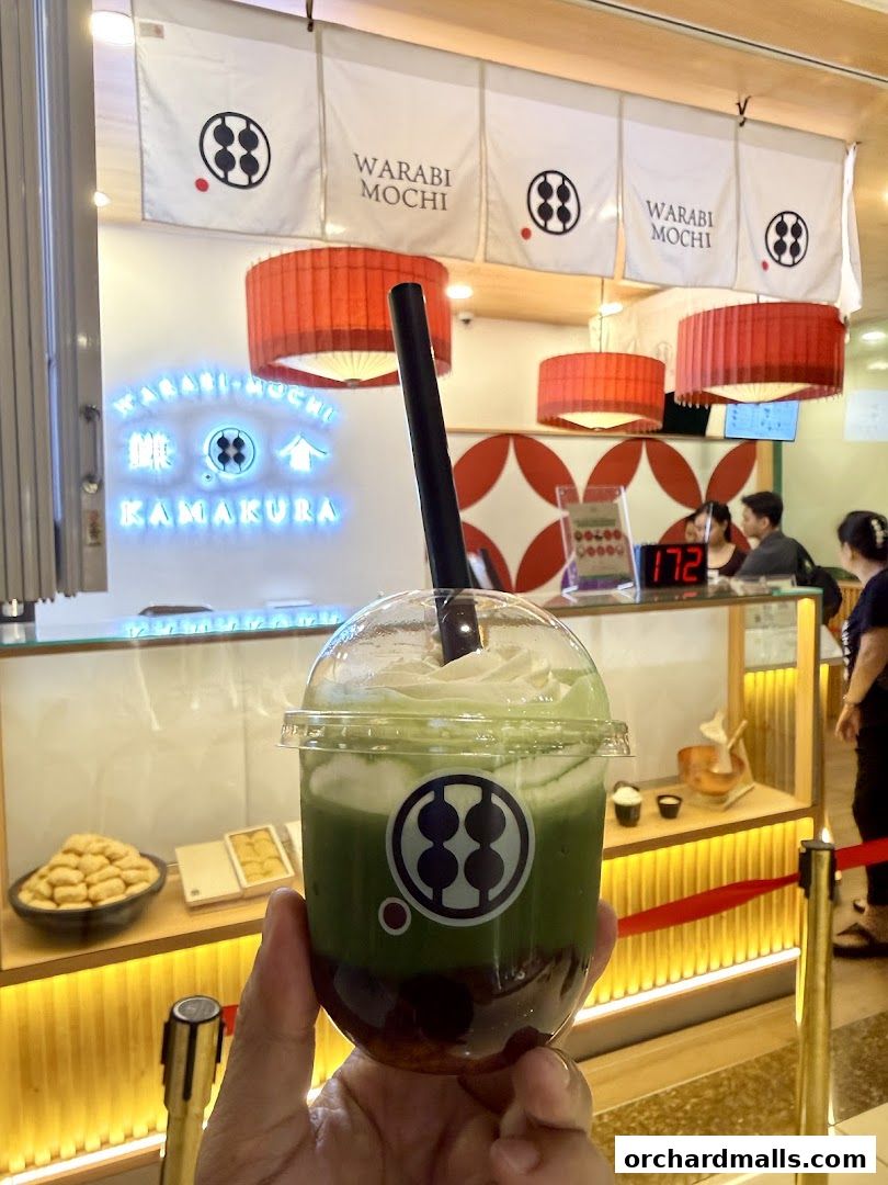 A hand holds a green matcha drink with whipped cream in front of a Warabi Mochi Kamakura shop.
