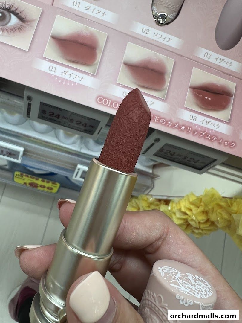 A hand holds a lipstick with a patterned bullet in front of a display of lipstick shades.