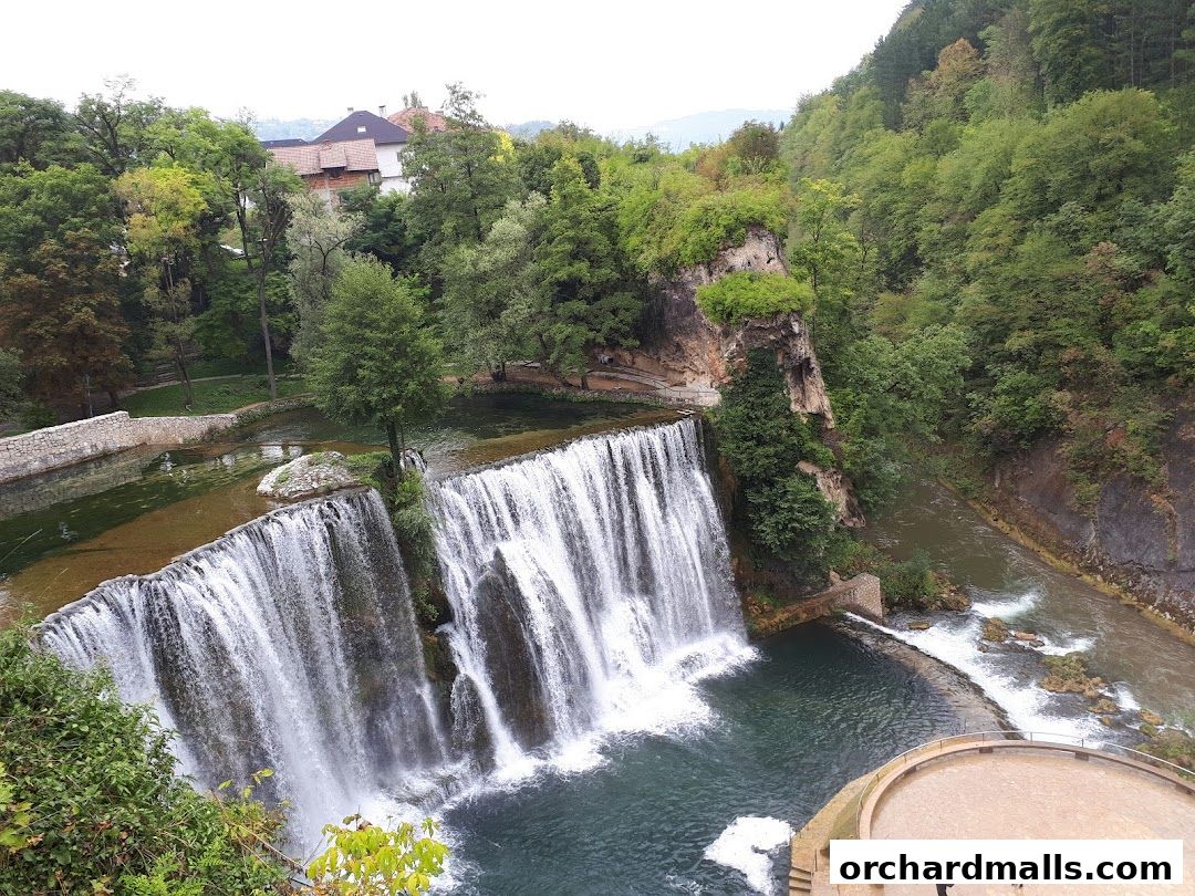 A wide waterfall cascades into a turquoise pool surrounded by lush green trees and rocky cliffs.