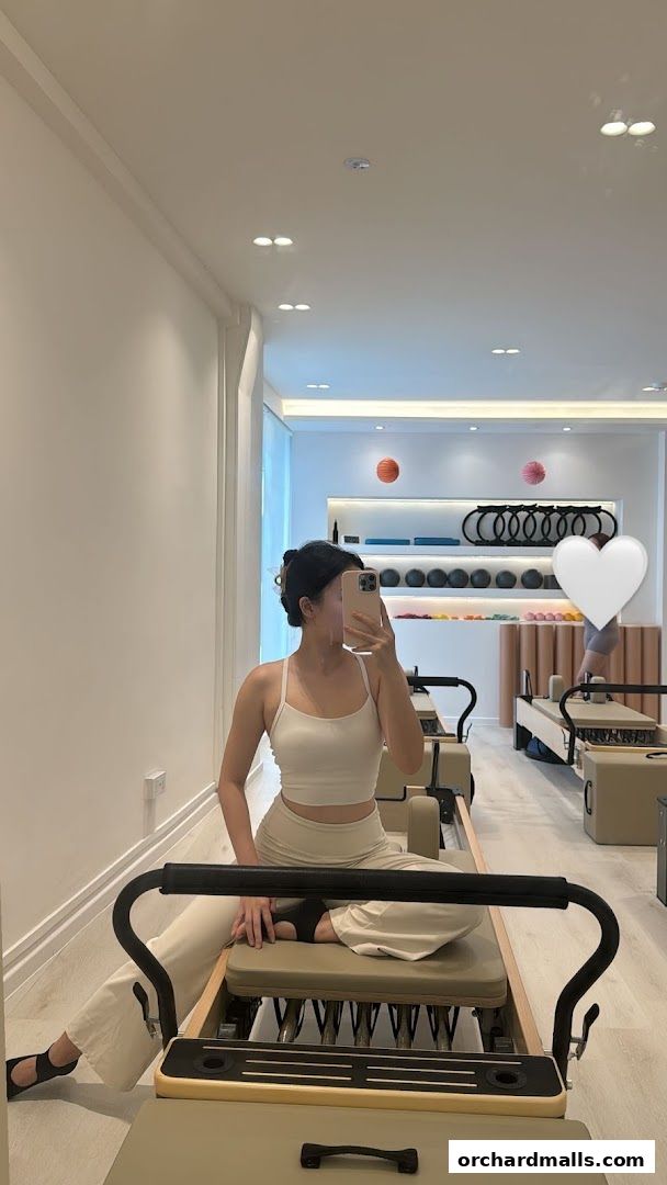 A woman in athletic wear sits on a Pilates reformer in a bright, modern studio.