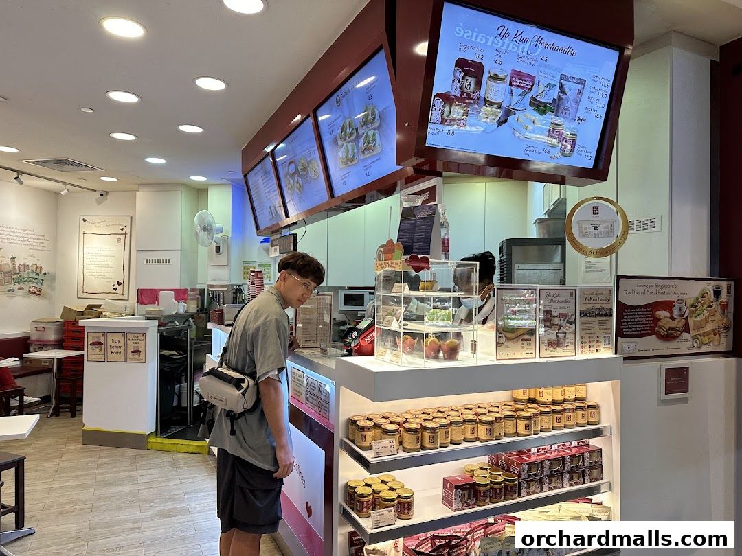 A customer stands at the counter of Ya Kun Kaya Toast, with merchandise and menu displays visible.