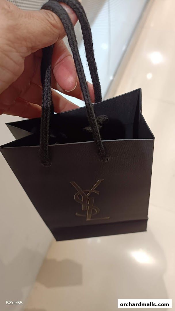 A hand holds a black YSL Yves Saint Laurent shopping bag with a gold logo.