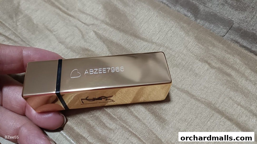 A hand holds a gold YSL lipstick with engraved text and logo.