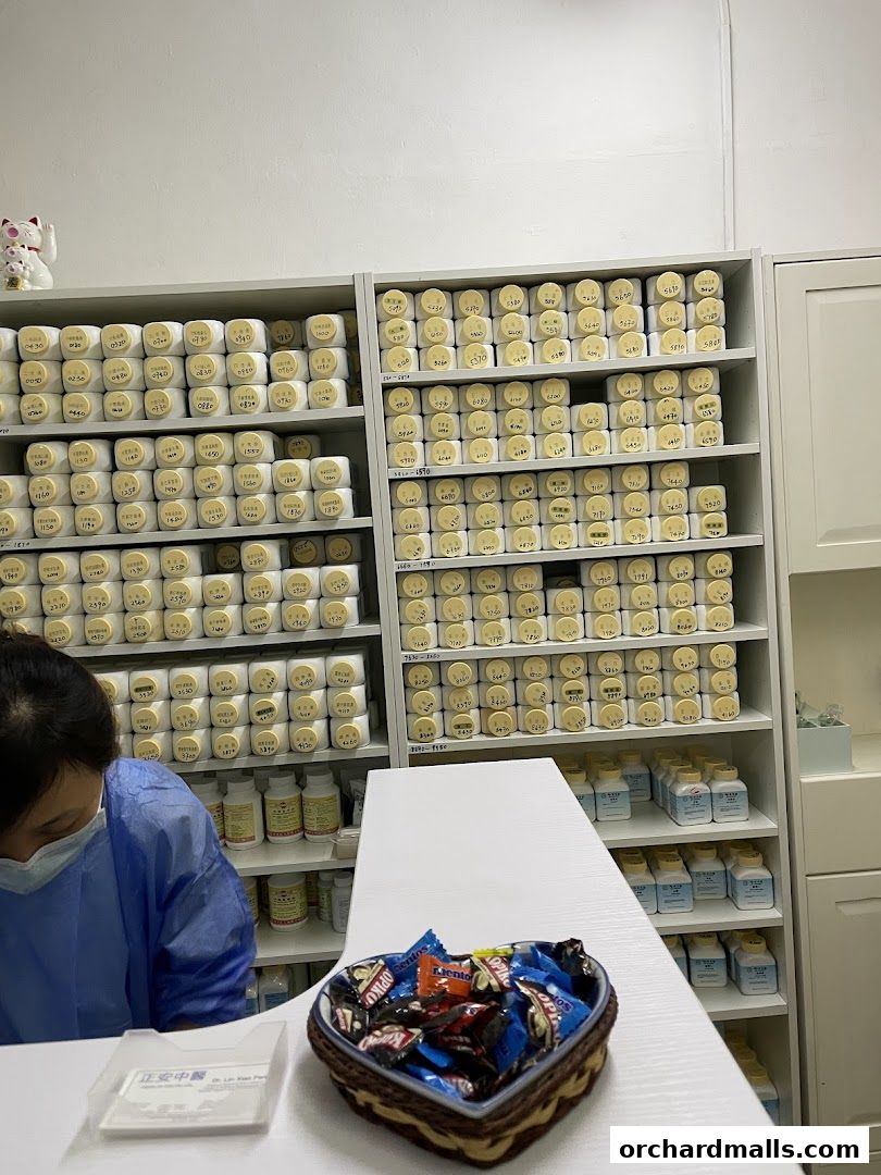A person in a blue gown works at a counter with shelves of traditional Chinese medicine bottles.