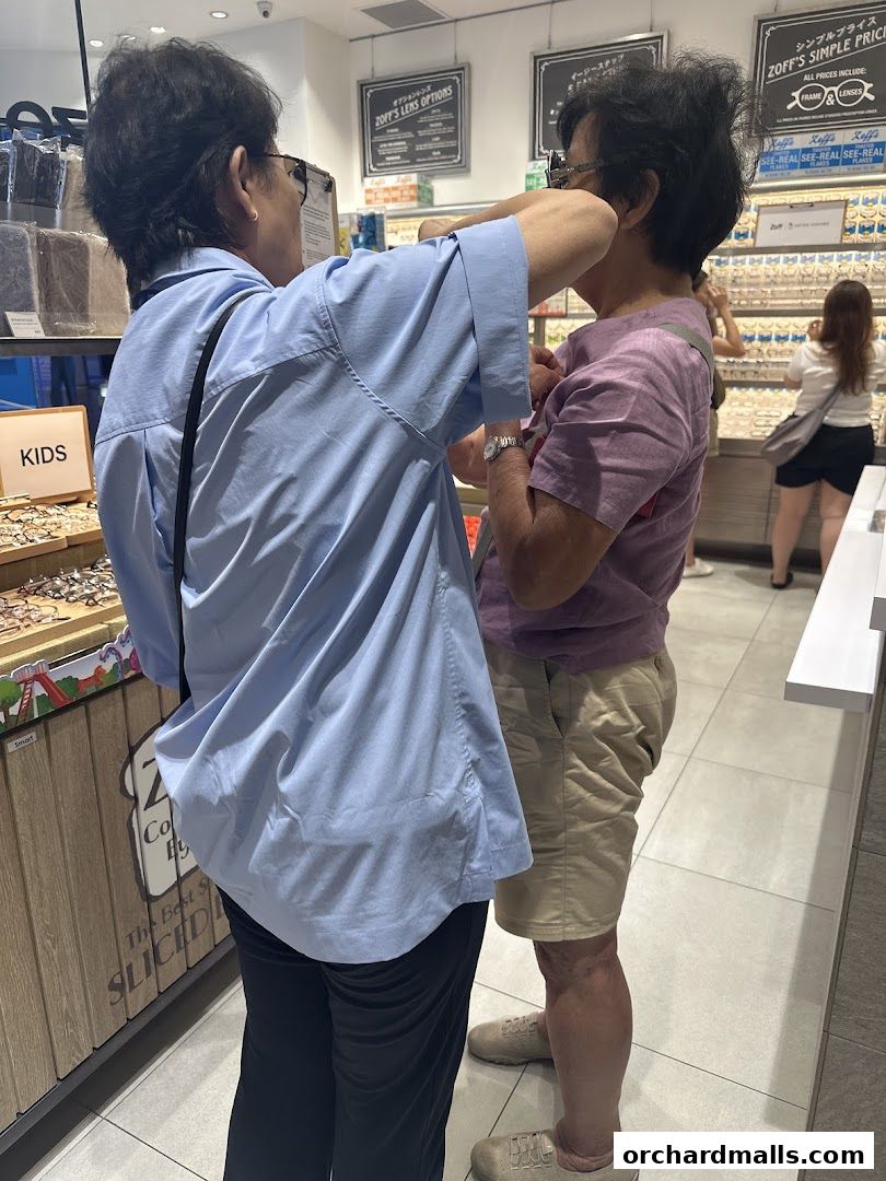 Two people are interacting inside an optical shop, with displays of glasses in the background.