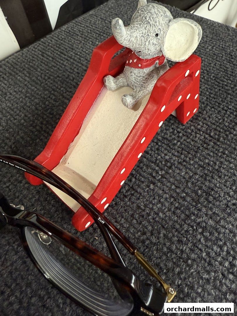 A pair of tortoiseshell glasses rests on a textured surface next to a decorative elephant on a red slide.